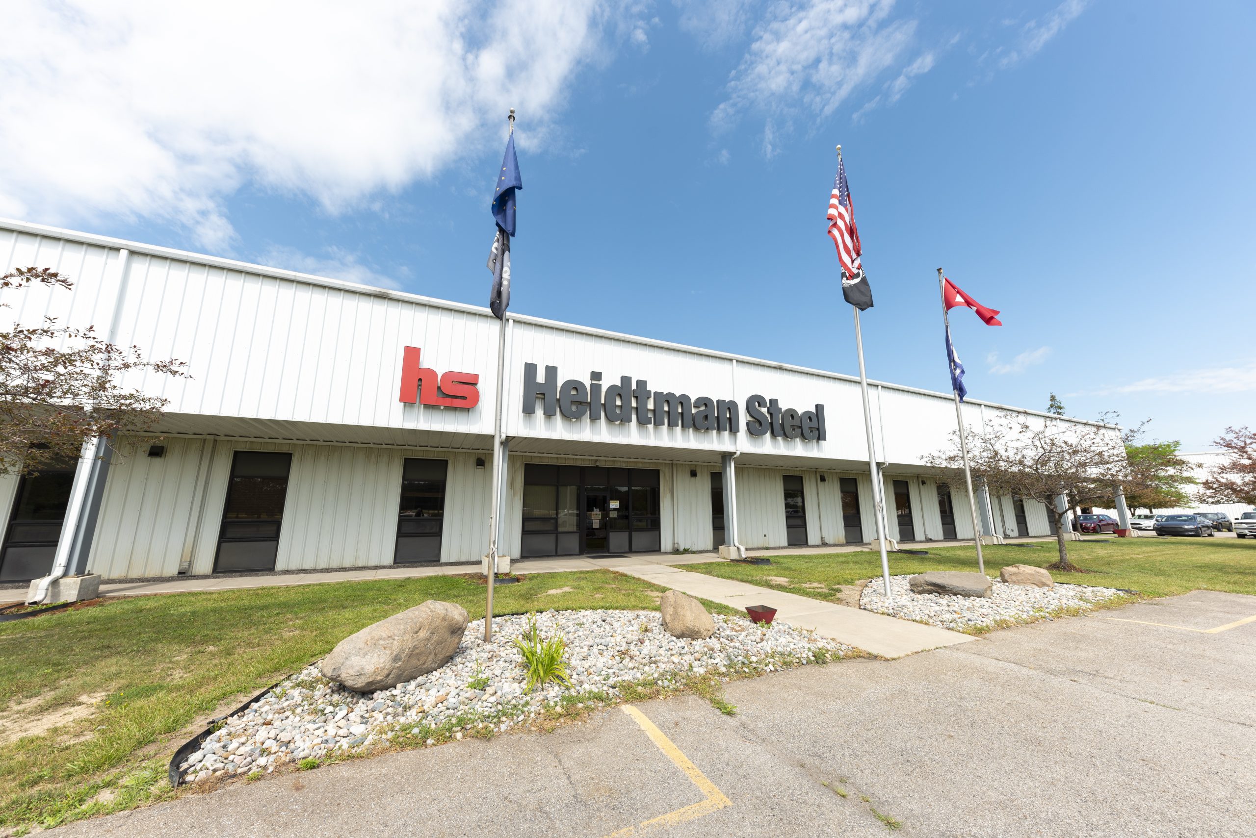 Heidtman Steel Butler, Indiana Facility Exterior White industrial building with Heidtman Steel signage, flags, parking lot, and rocks under a blue sky.