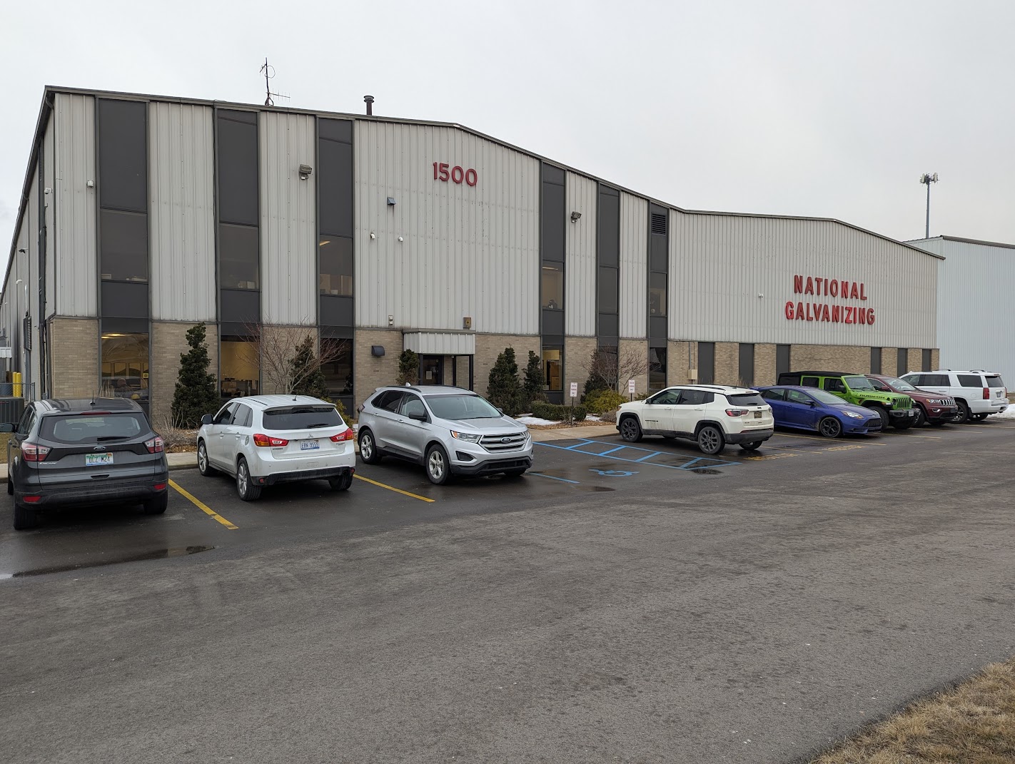 National Galvanizing Facility Exterior Gray industrial building with 'NATIONAL GALVANIZING' and '1500' signage, parking lot, and cars on a cloudy day.